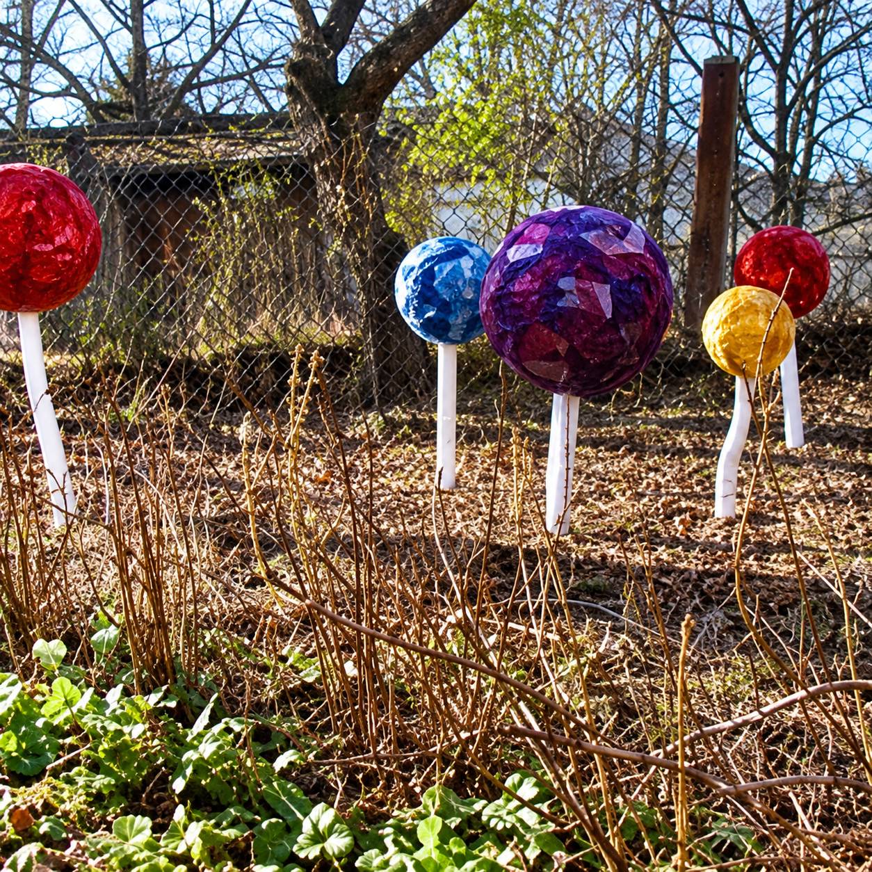 Des boules décoratives en verre rouges, bleues, violettes et jaunes sont posées sur de fines tiges blanches dans un jardin aux herbes sèches.