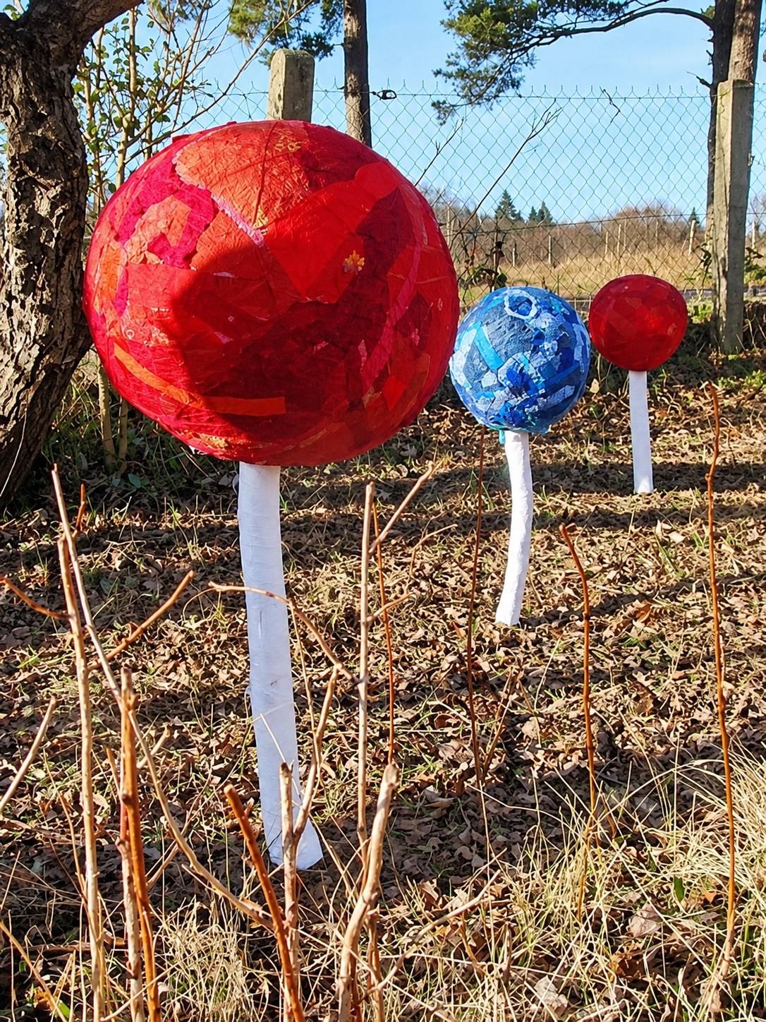 Trois grands champignons décoratifs, un rouge, un bleu et un petit rouge, plantés sur des pieds blancs dans l'herbe sèche d'un jardin.
