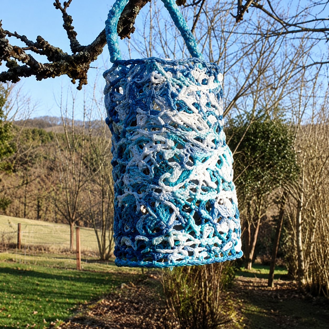 Une lanterne cylindrique ajourée aux motifs bleus et blancs est suspendue à une branche, devant un paysage de collines ensoleillées.