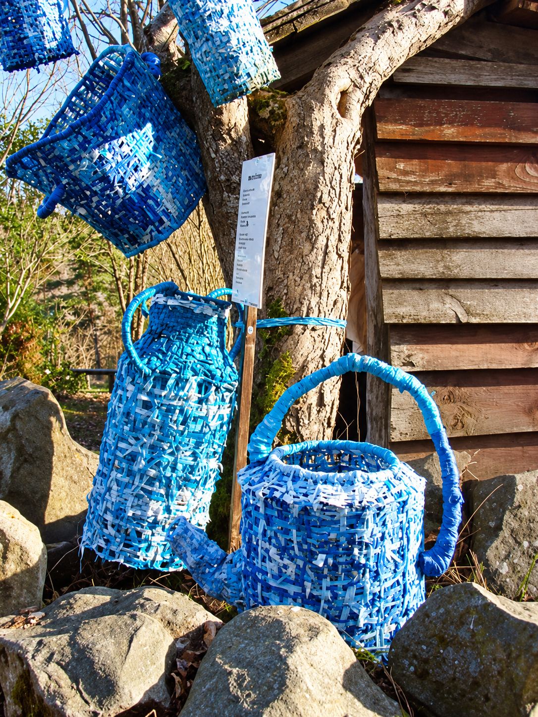 Des sculptures tressées bleues, dont un arrosoir et un pichet, sont exposées sur des rochers devant un arbre et un mur en bois, avec des paniers suspendus.