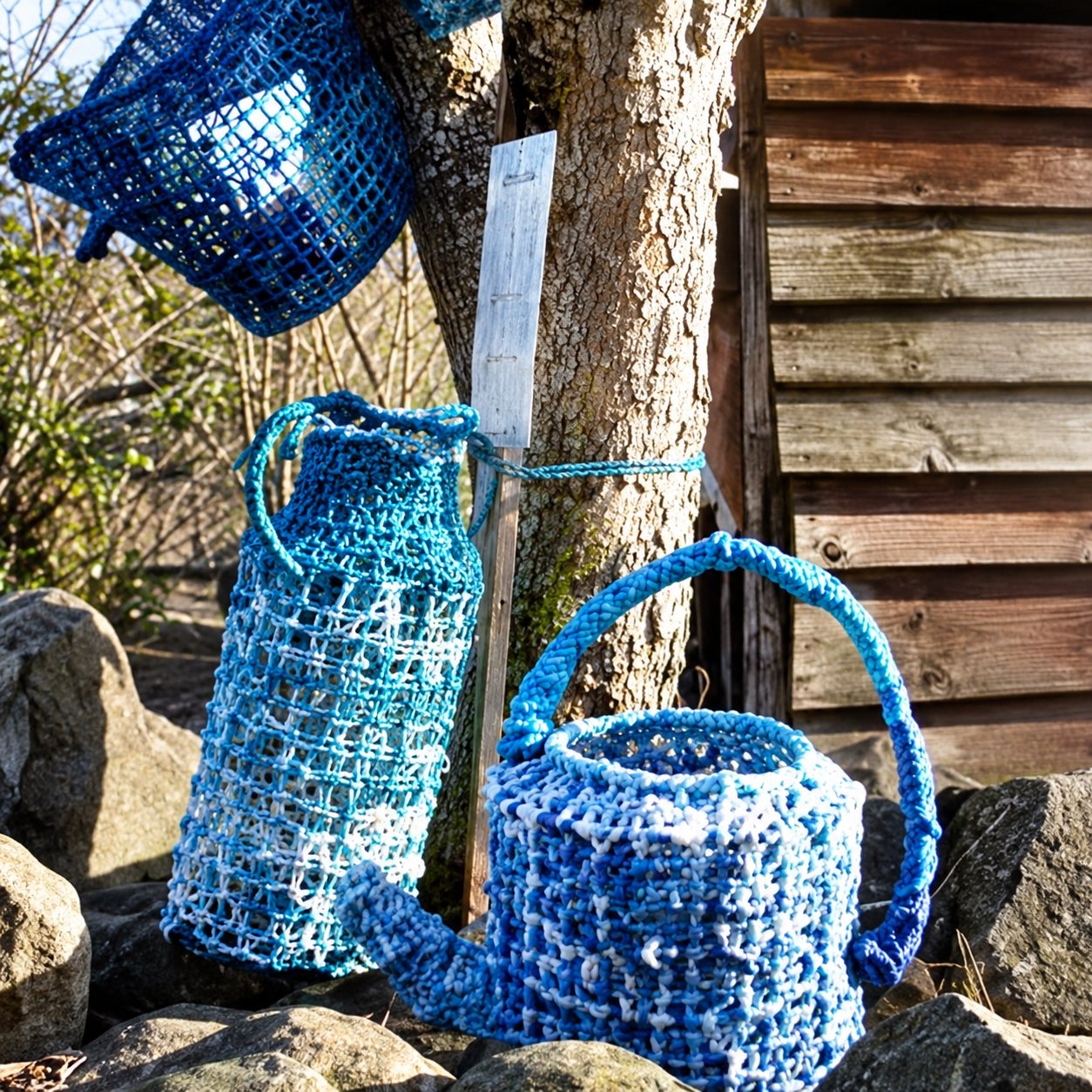 Deux paniers en maille bleue à anses sont posés à l'extérieur près d'un tronc d'arbre, devant un mur en bois, avec un autre objet bleu suspendu en haut à gauche.