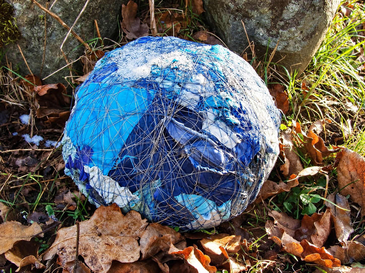 Une grosse boule bleue et blanche enveloppée d'un filet est posée dans l'herbe parmi des feuilles mortes, au pied d'une pierre.